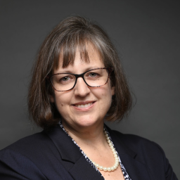 Headshot for Katie Brisson, smiling towards the camera wearing a suit jacket and glasses